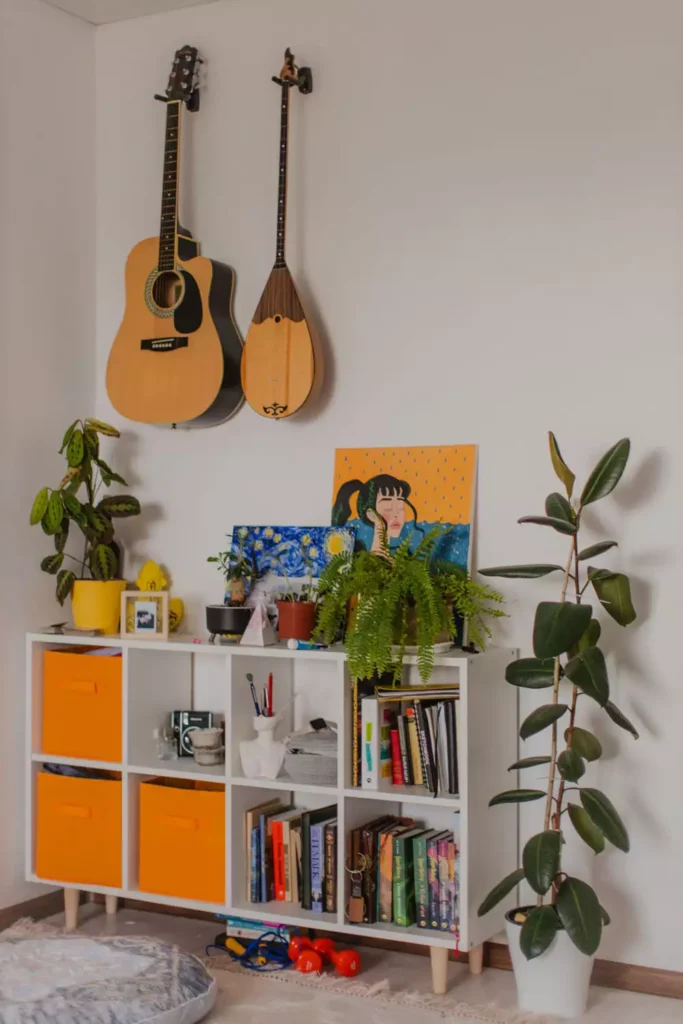 A guitar and a saz-style stringed instrument used as wall decoration above a colorful bookshelf and plants