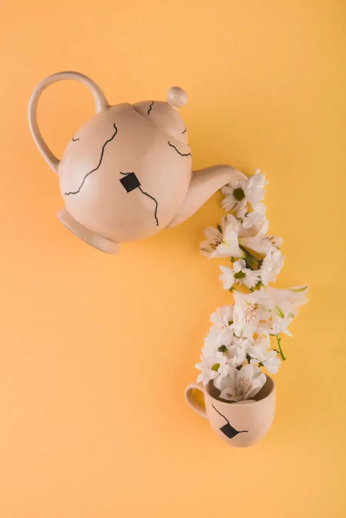 A cream ceramic teapot with black line details pours white flowers into a matching cup, styled as a ceramic wall decor still life on yellow