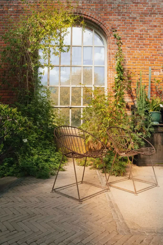 Arched Window On An Outdoor Brick Wall Framed By Climbing Greenery And Iron Chairs