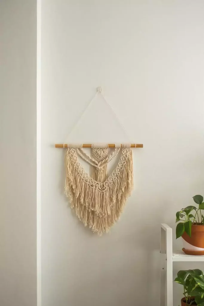 Boho macrame wall hanging on a wooden dowel beside a white shelf with potted plants for easy aesthetic DIY room decor