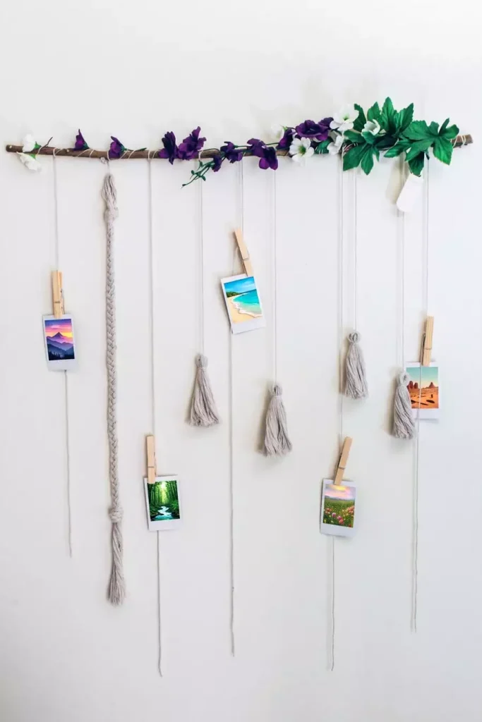 Branch wall hanger with faux flowers yarn tassels and wooden clothespins holding small landscape photos as easy aesthetic DIY room decor