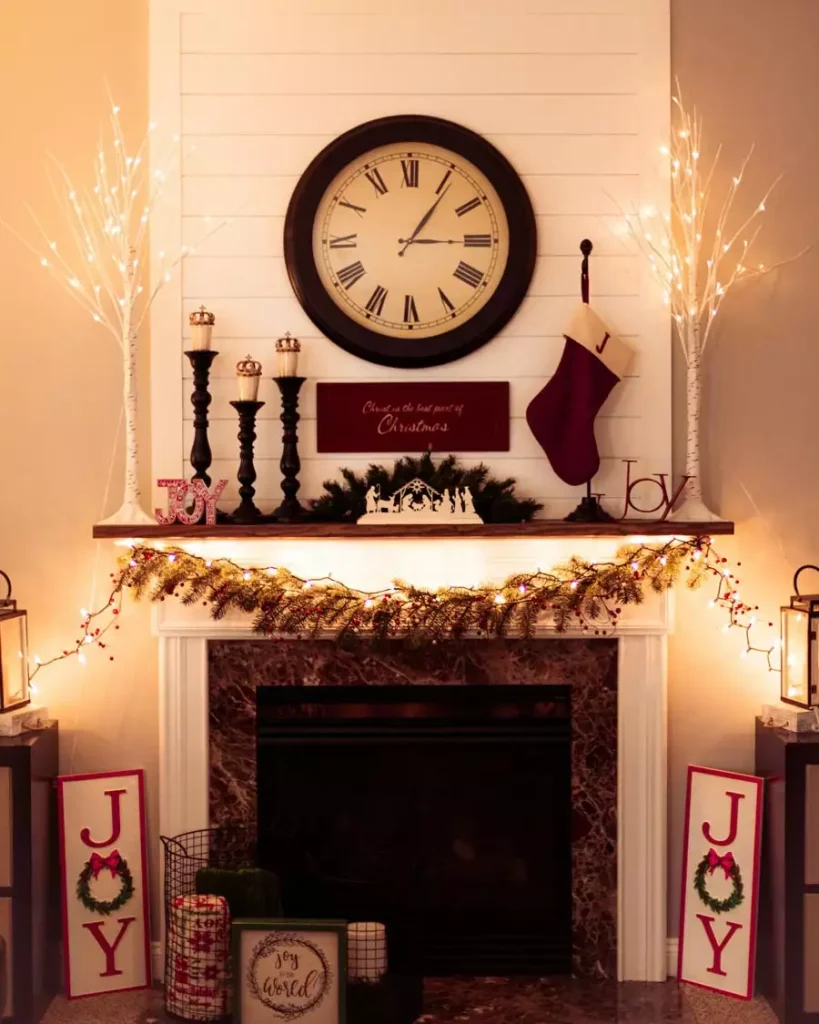 Christmas Wall Mantel Styled With A Round Clock, Joy Signage, A Stocking, And Lit Birch Branches