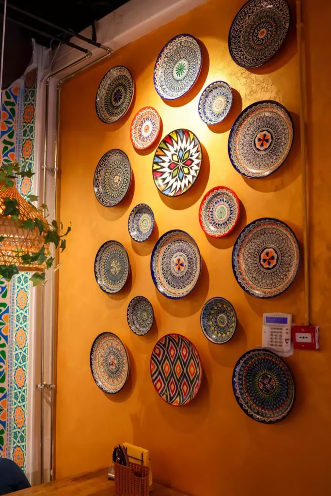 Colorful mandala and diamond plates are spaced across a mustard restaurant wall as ceramic wall decor under warm indoor lighting