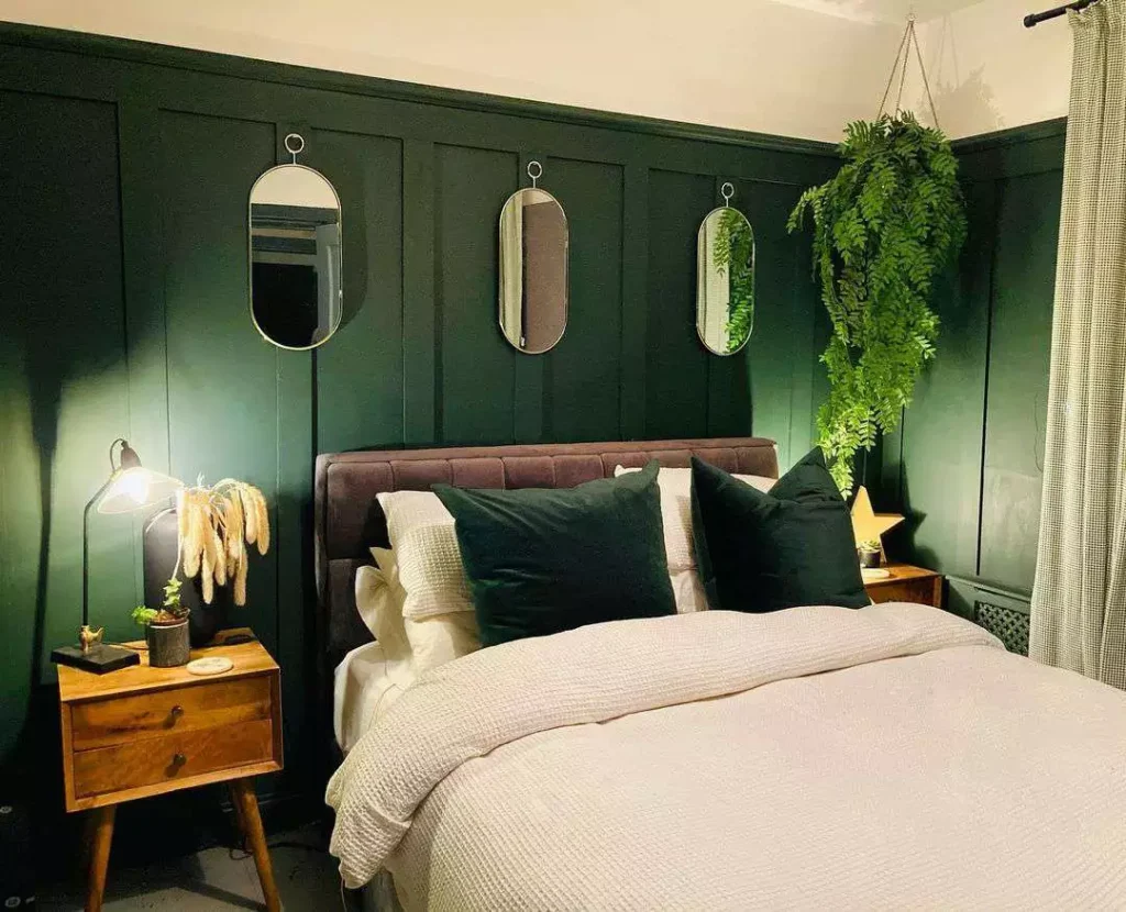 Dark green wall bedroom with paneled paint, oval mirrors, hanging fern, and cream bedding