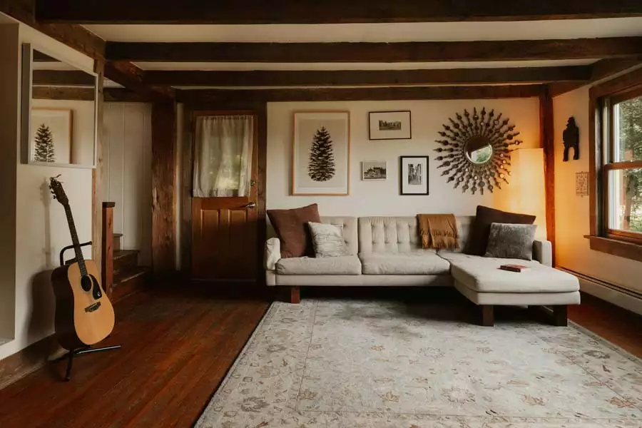 Exposed-Beam Living Room Farmhouse Wall Featuring A Pinecone Print, Small Framed Art, And A Sunburst Mirror Above A Light Sectional Sofa