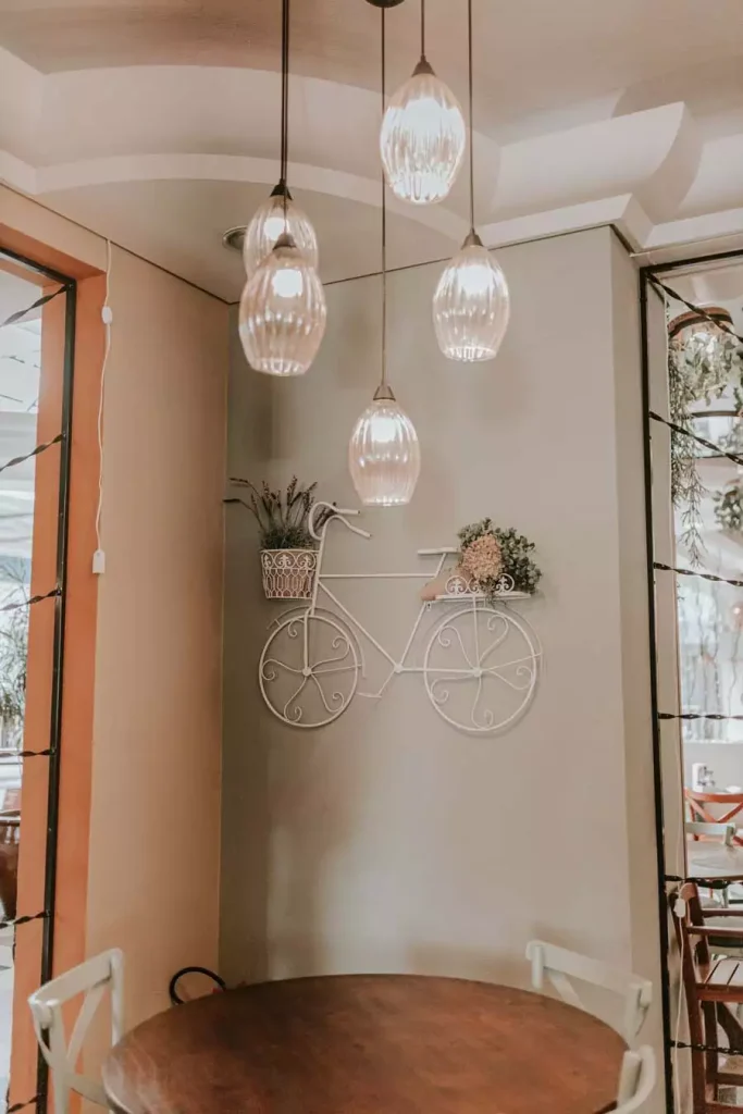 Farmhouse Wall Accent Piece Shaped Like A Wire-Frame Bicycle With Faux Flower Baskets, Mounted Beneath Hanging Glass Pendant Lights Above A Café Table