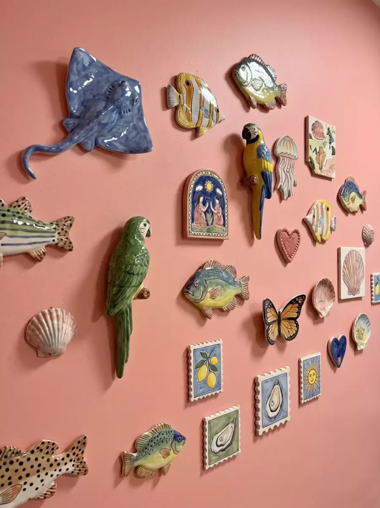Glazed stingray, parrot, fish, shell, butterfly, and tile pieces cover a pink wall as playful ceramic wall decor in a dense mixed gallery