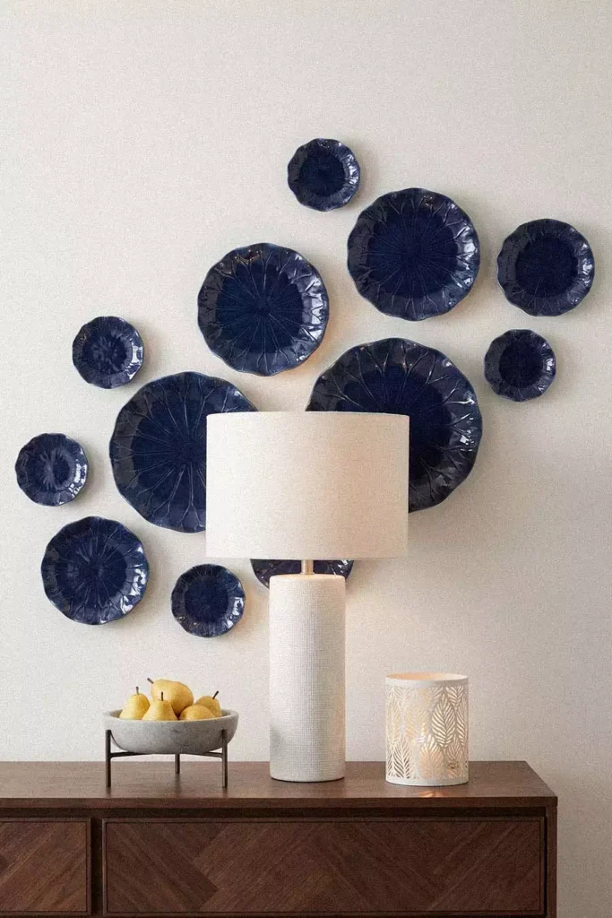 Glossy navy lotus leaf plates cluster behind a white drum lamp as ceramic wall decor above a herringbone wood dresser with pears and a pierced lantern