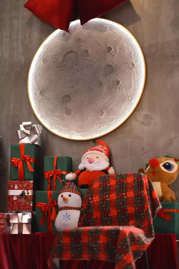 Glowing Moon Christmas Wall Art Above Wrapped Gifts, A Plush Santa, And A Snowman