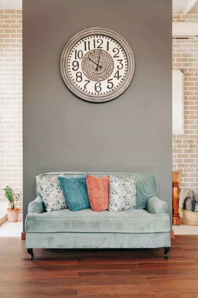 Large Ornate Round Wall Clock Centered On A Charcoal-Gray Wall Above A Sage-Green Loveseat With Mixed Throw Pillows