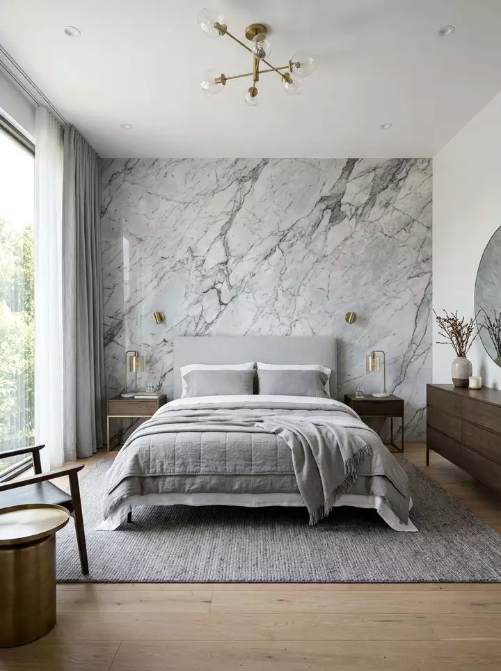 Light grey marble bedroom wall with brass sconces and a round mirror over a wood dresser