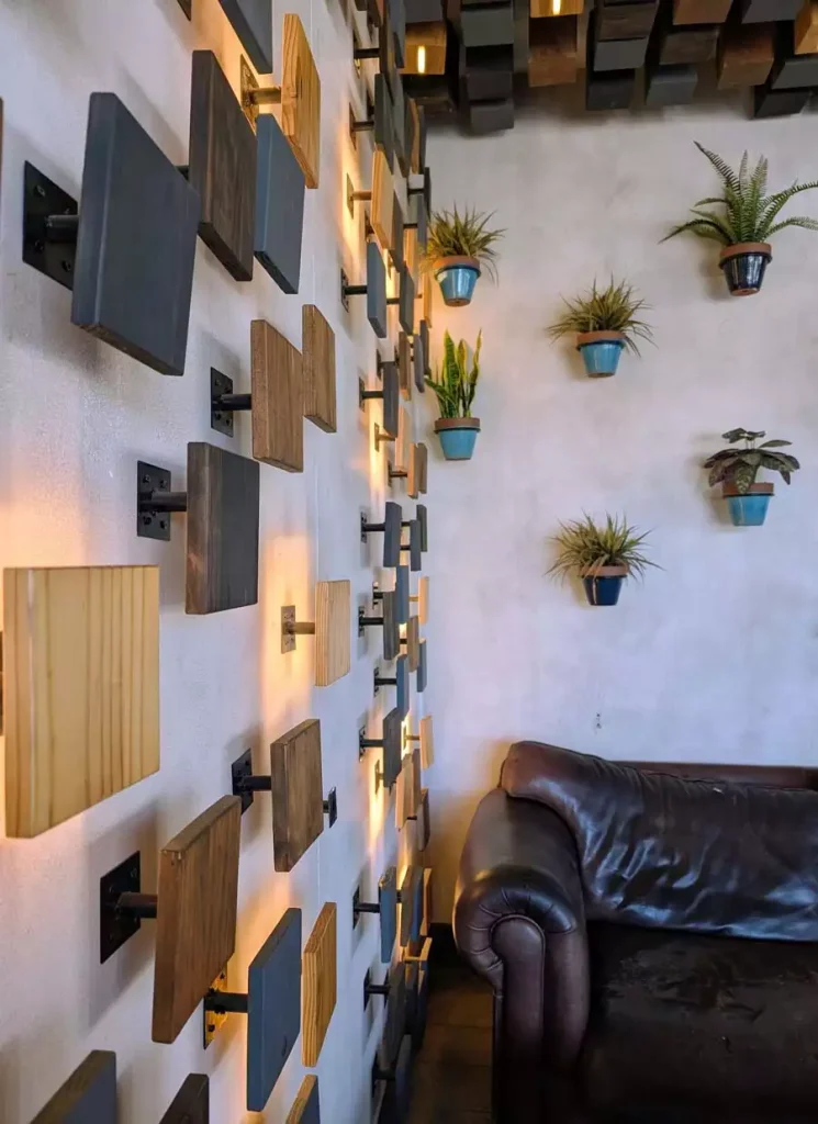 Modern Accent Wall With Layered Wooden Wall Decor Blocks, Warm Sconce Lighting And Hanging Potted Plants