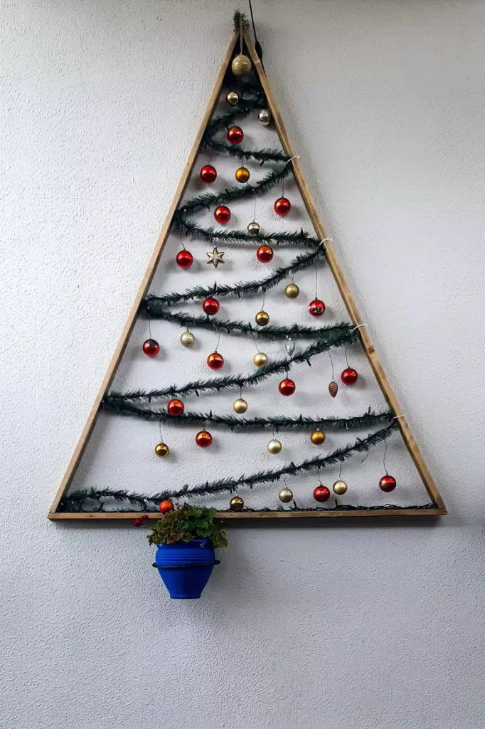Outdoor Triangular Christmas Wall Tree Frame Strung With Lights And Ornaments Over A Blue Planter