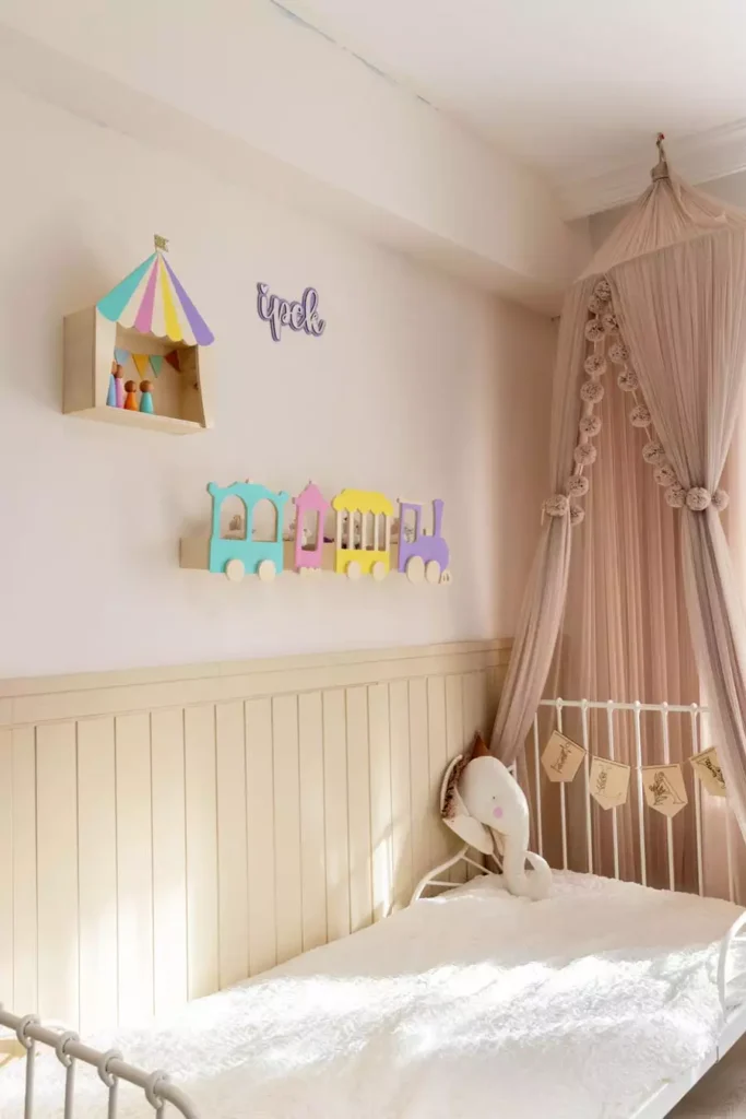 Pastel circus tent shelf and train shaped ledge with a name sign, colorful nursery wall decor beside a blush canopy bed