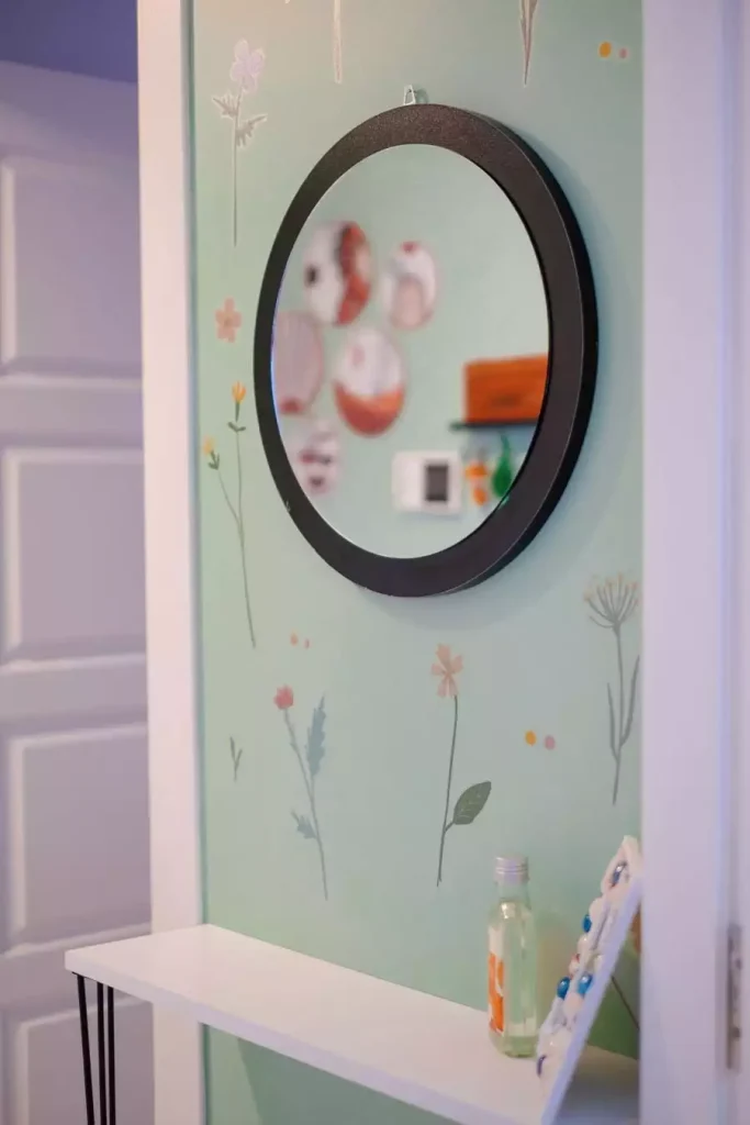 Round black mirror on a mint green wall with delicate wildflower illustrations above a white shelf for easy aesthetic DIY room decor