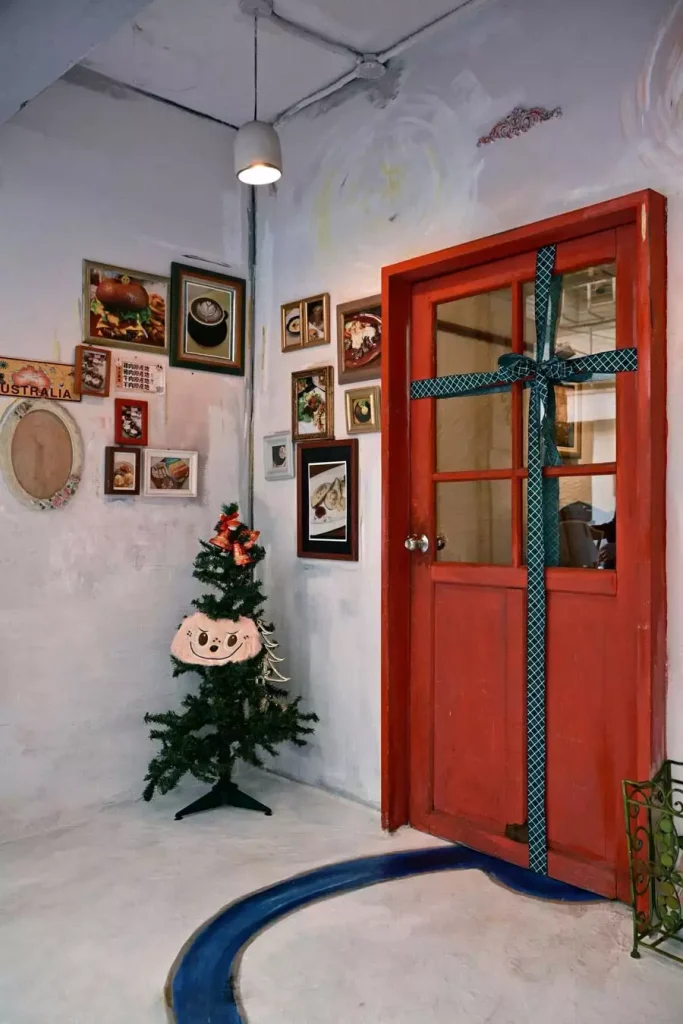 Small Christmas Tree In A Loft Corner Beside A Gift-Wrapped Red Door And A Photo Wall