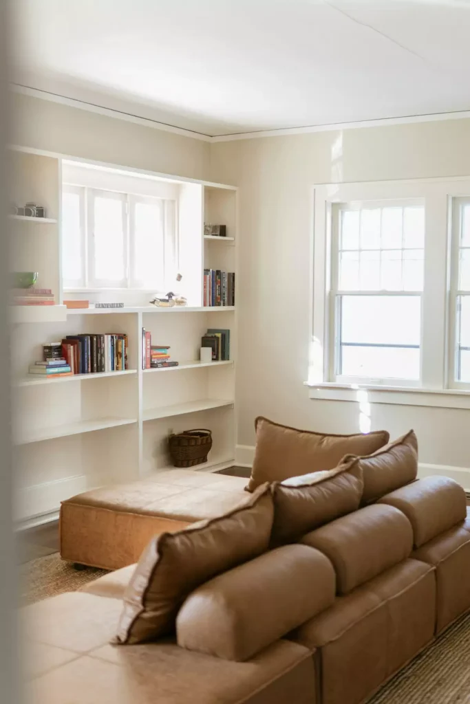 Sunlit tan wall living room reading nook with a camel leather sectional and white built-in bookshelves