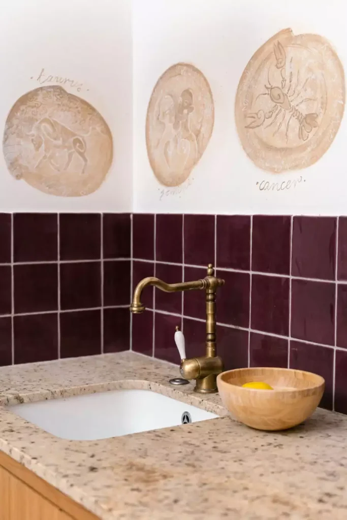Taurus, Gemini, and Cancer roundels sit above burgundy square tiles as ceramic wall decor in a kitchen with a brass faucet and stone counter
