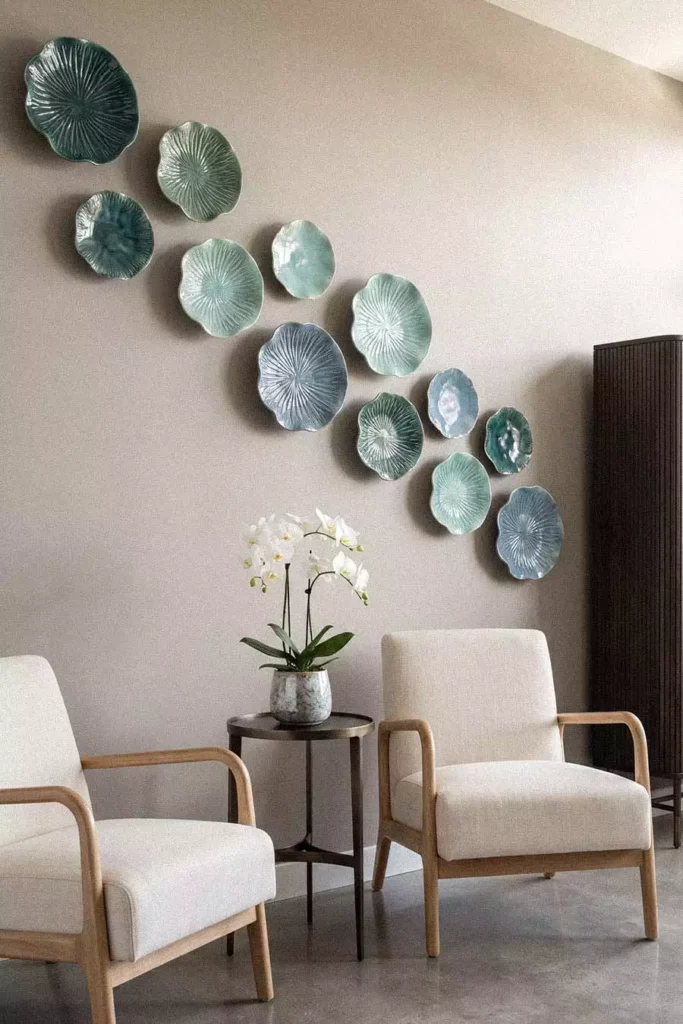 Teal glazed lotus leaf plates sweep across a taupe wall as ceramic wall decor above two cream wood armchairs, a round metal side table, and a white orchid
