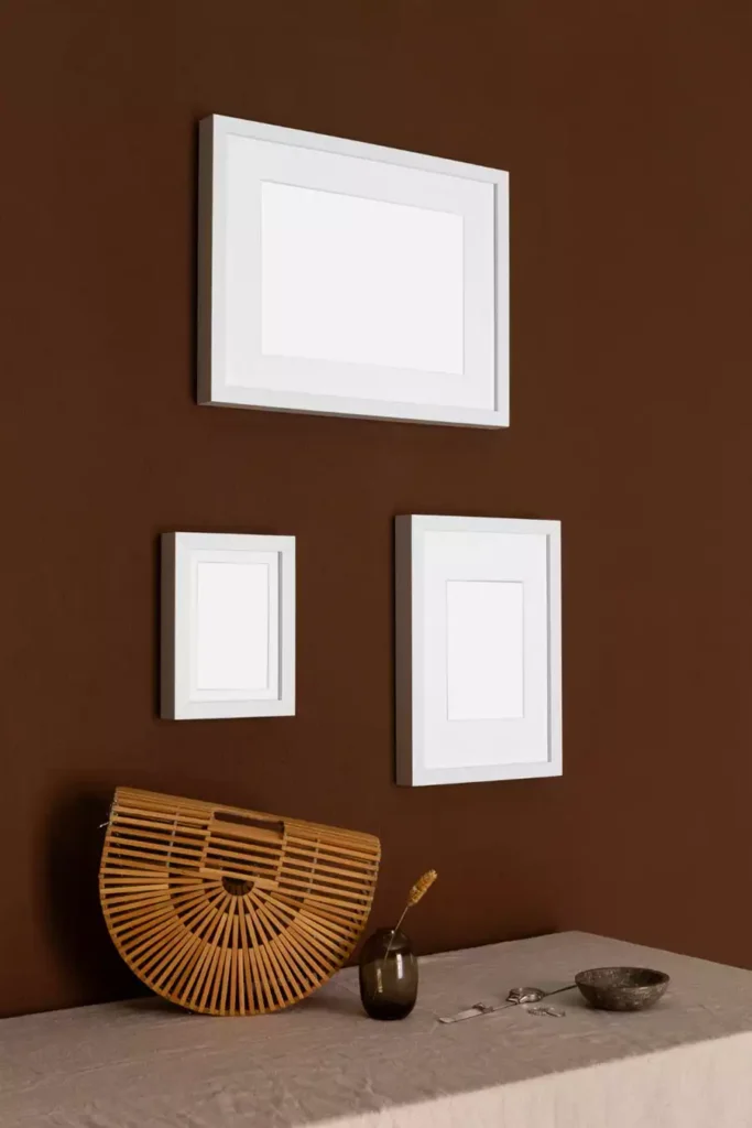 Three empty white frames hang on a chocolate brown wall in a simple gallery layout that can sit with ceramic wall decor styling below