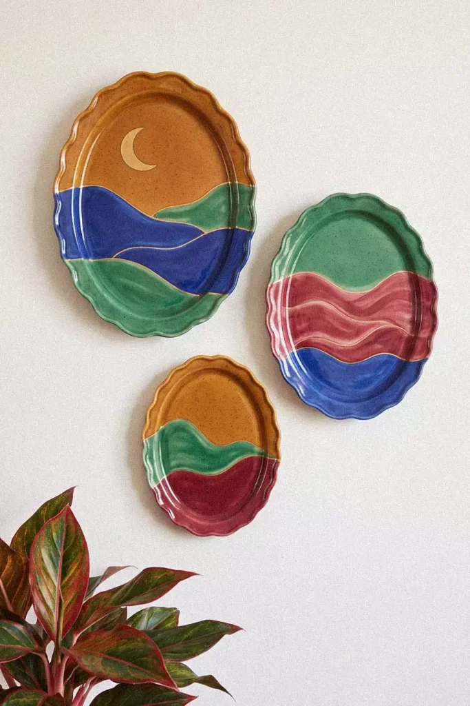 Three scalloped oval plates painted with hills, waves, and a crescent moon form ceramic wall decor beside a red and green houseplant