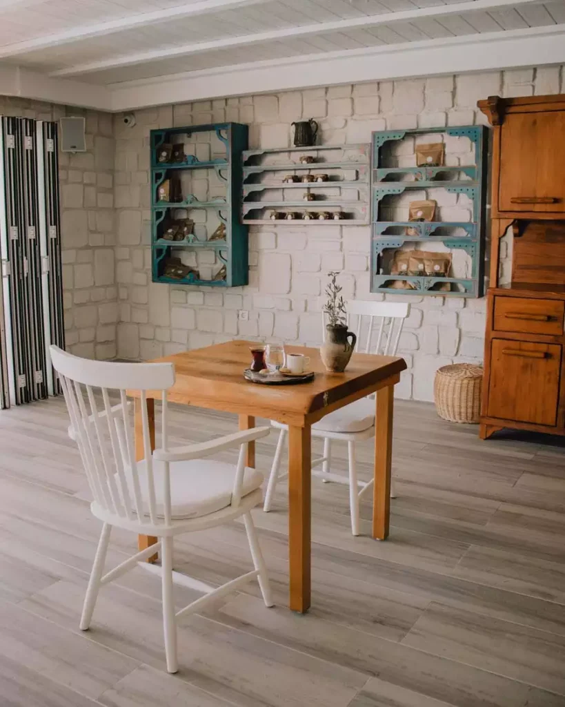 Whitewashed Stone Farmhouse Wall Fitted With Teal Wall-Mounted Shelving Units Stocked With Packaged Goods Above A Wooden Dining Table