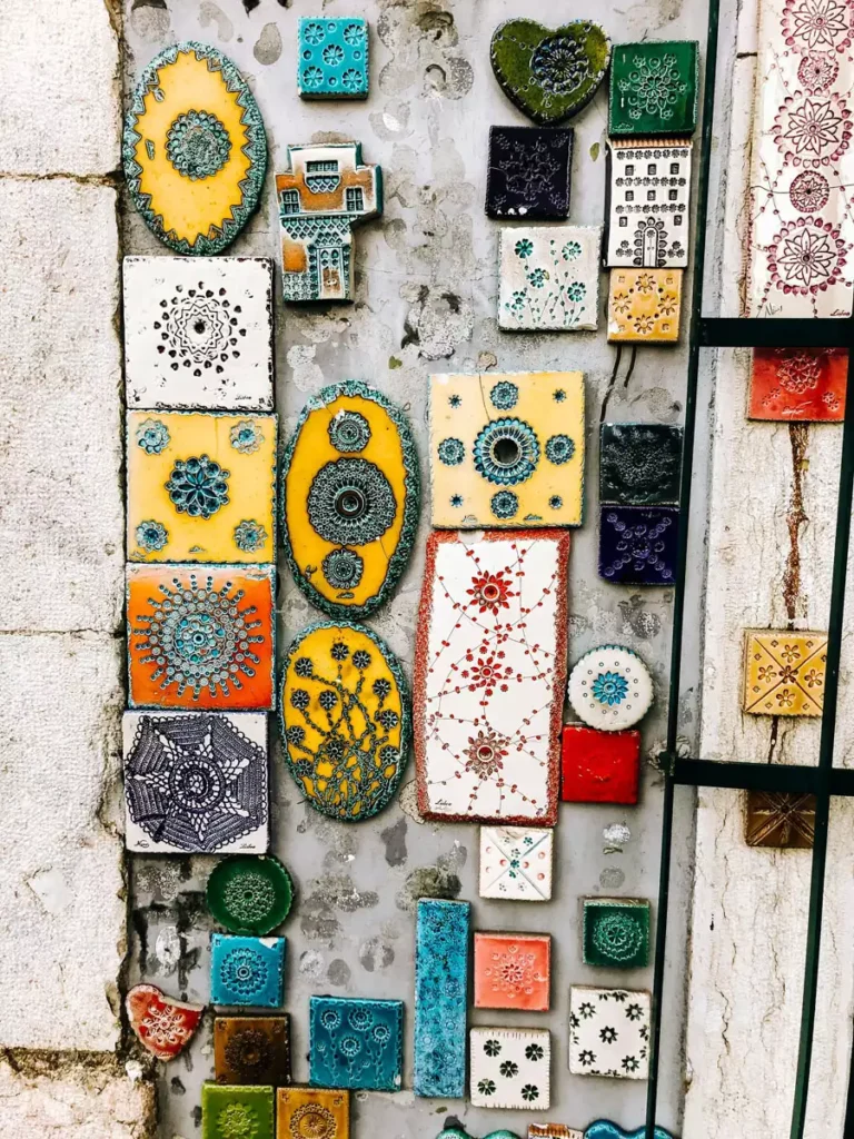 Yellow, teal, terracotta, and white handmade tiles of many shapes cover a weathered wall as ceramic wall decor next to a black metal grille