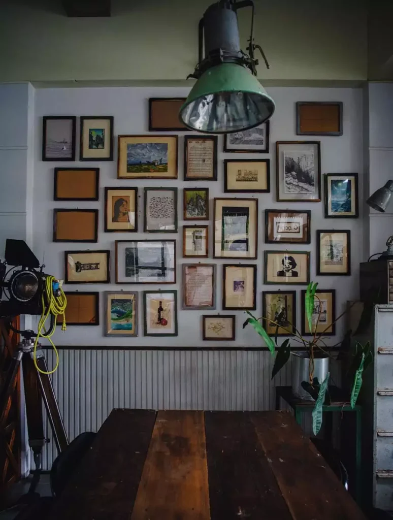 A dense collection of mixed-size framed artwork fills the wall above paneling and a wooden table beneath a green industrial pendant light