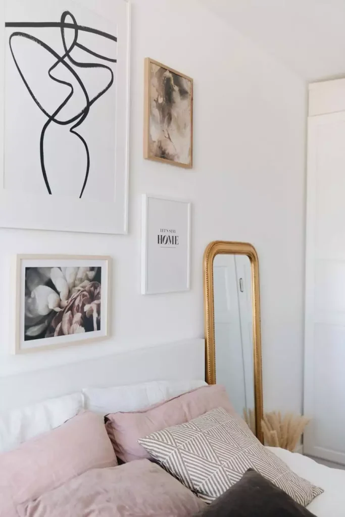 A large abstract line drawing and smaller floral and text prints hang above a bed with pink pillows beside a tall gold-framed mirror
