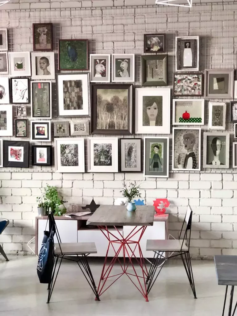 A tightly packed collection of framed portraits, drawings, and paintings covers a white brick wall above a small table and chairs