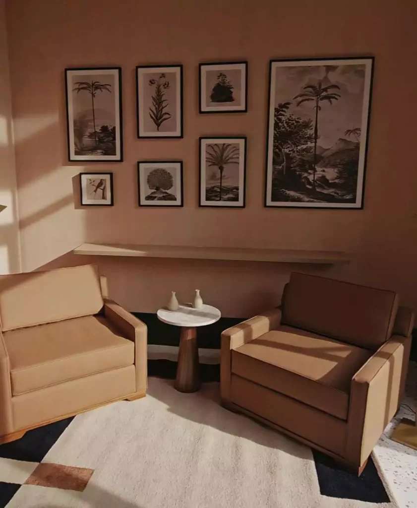 Botanical gallery wall decor features mixed-size black frames above two tan armchairs and a small round table against a peach wall