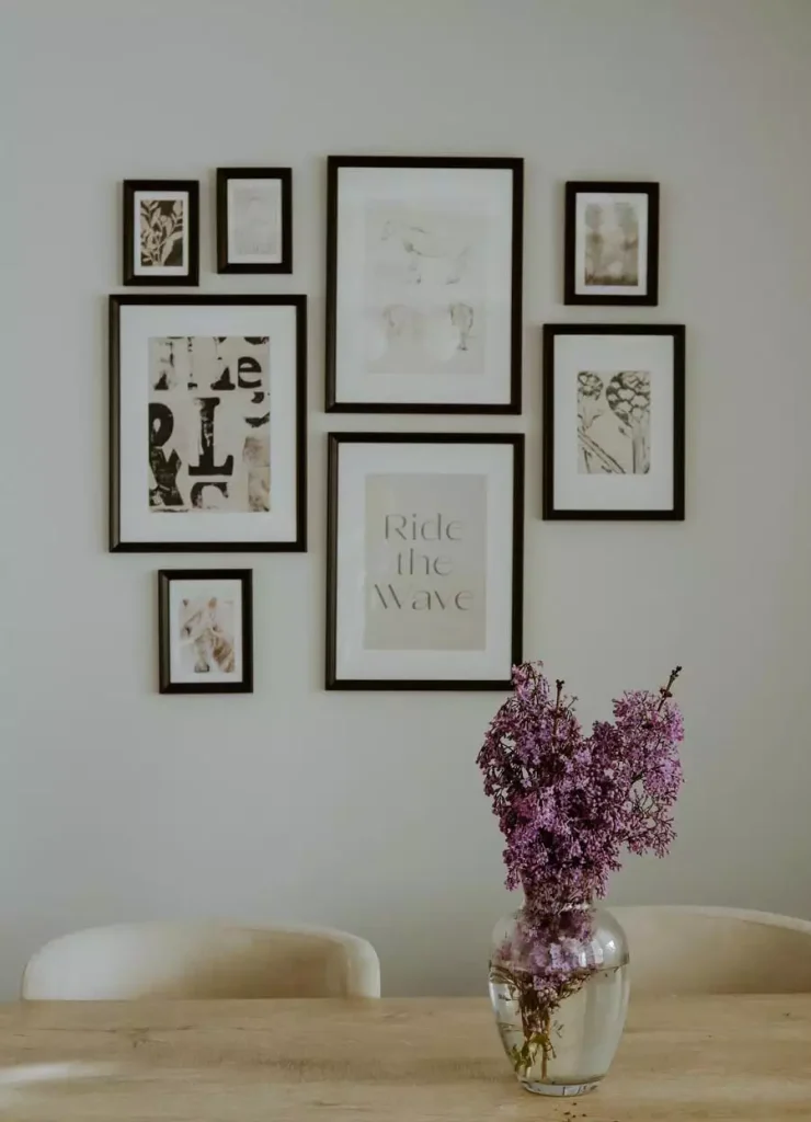 Eight black frames display pale illustrations, botanical prints, and typography above a wooden dining table with purple flowers