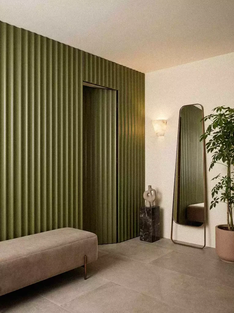 Entryway with olive-green fluted wall panels, concealed door, taupe upholstered bench, bronze-framed mirror, and Schefflera plant