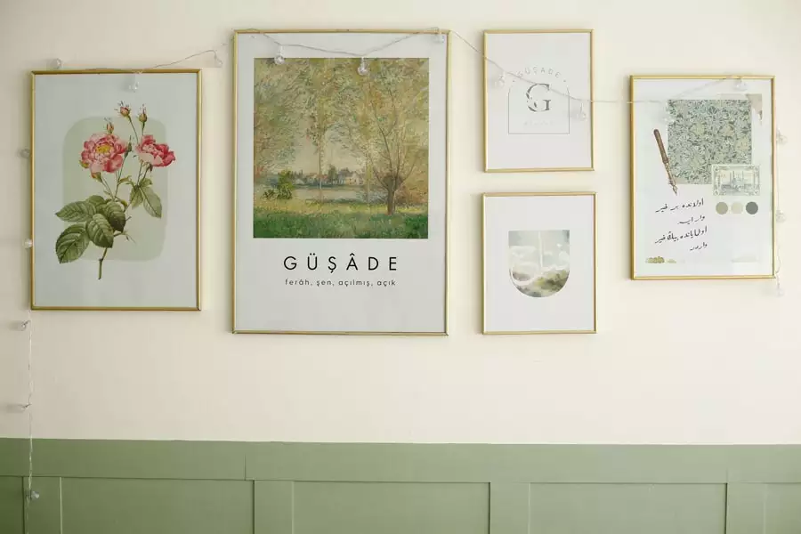 Five thin gold frames display floral, landscape, and minimalist prints, with string lights draped above sage-green wall paneling