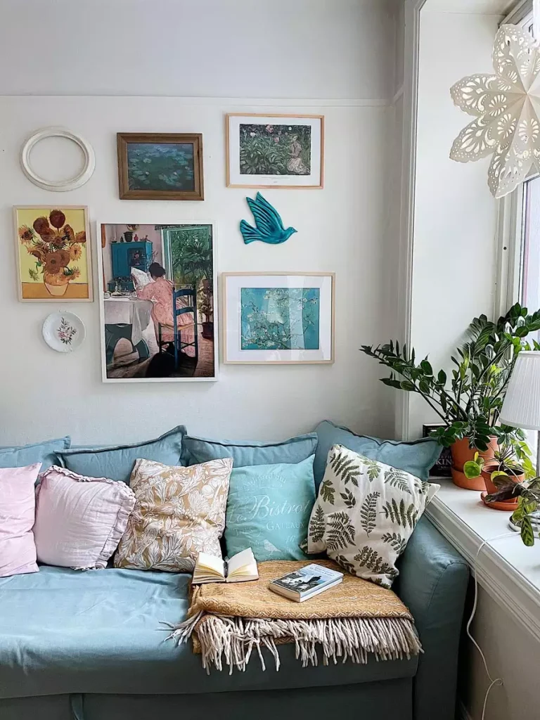 Framed paintings, decorative plates, and a turquoise bird ornament hang above a pale blue sofa beside a window filled with plants