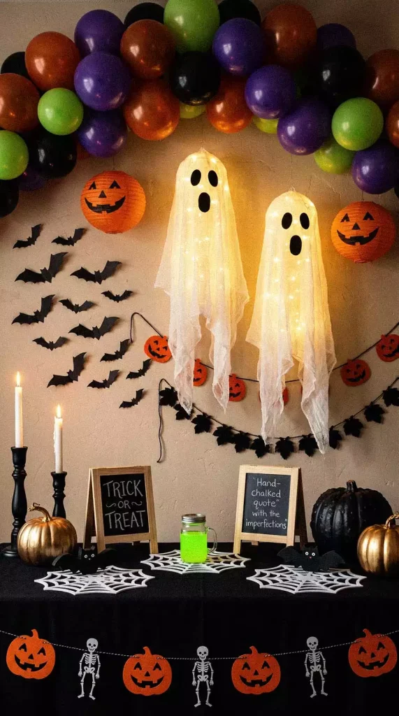 Glowing hanging ghosts beneath a colorful balloon arch, with bats and garlands above a Halloween party table