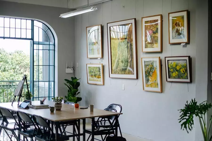 Gold-framed landscape and floral paintings hang above a long table with black chairs beside a tall arched window