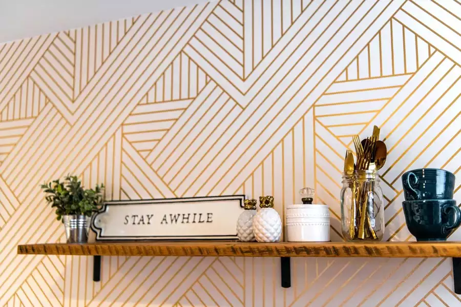 Gold geometric wallpaper behind a wooden kitchen shelf with a Stay Awhile sign