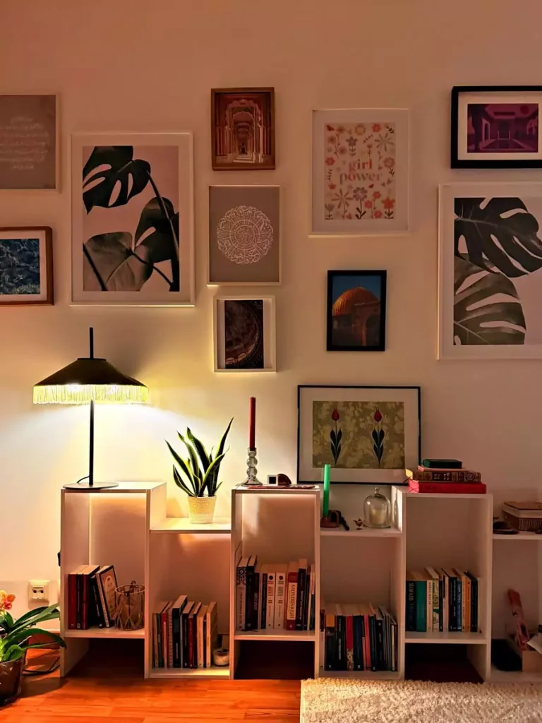 Leaf prints, floral illustrations, and smaller framed artworks hang above a white bookcase filled with books beneath warm lamp lighting