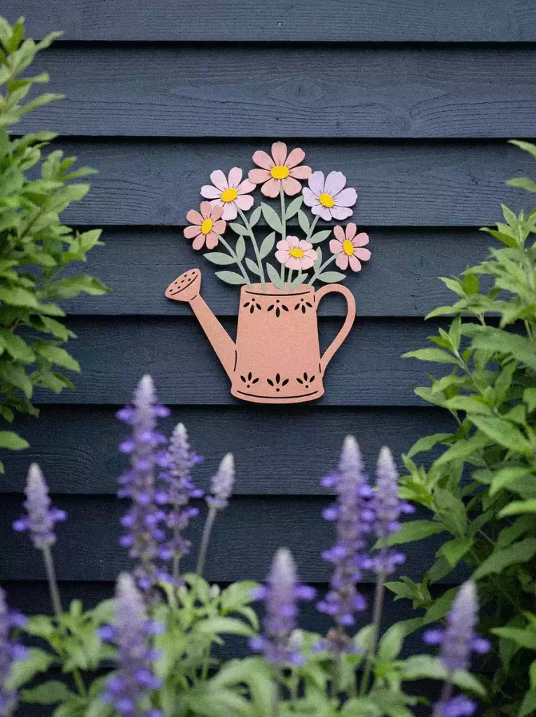 Metal watering-can wall art with pink and lilac flowers on dark navy wooden siding, framed by garden foliage