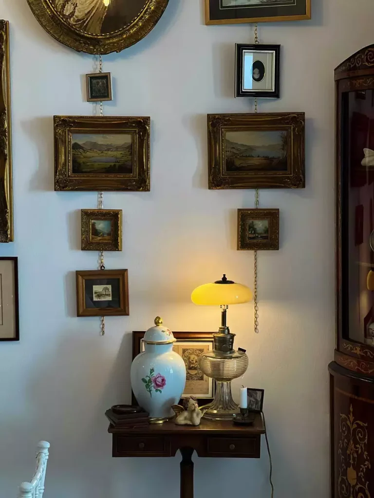 Ornate landscape paintings and smaller framed photographs hang from vertical chains above a wooden console with a yellow lamp and floral vase