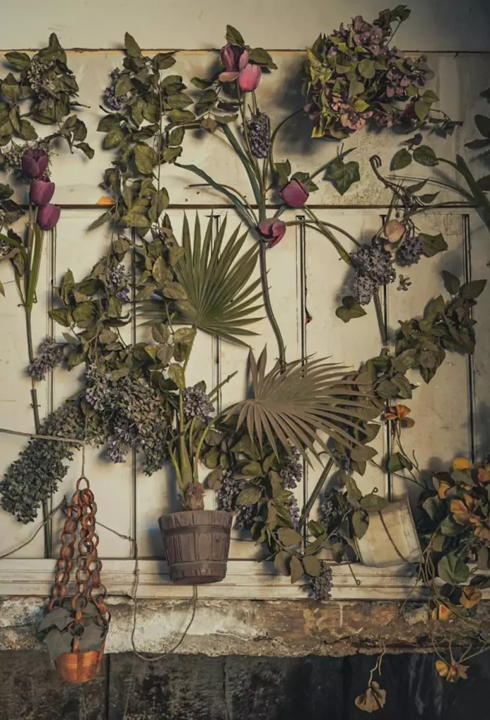 Rustic floral wall display with pink flowers, trailing foliage, fan-shaped leaves, and hanging pots against cream panels