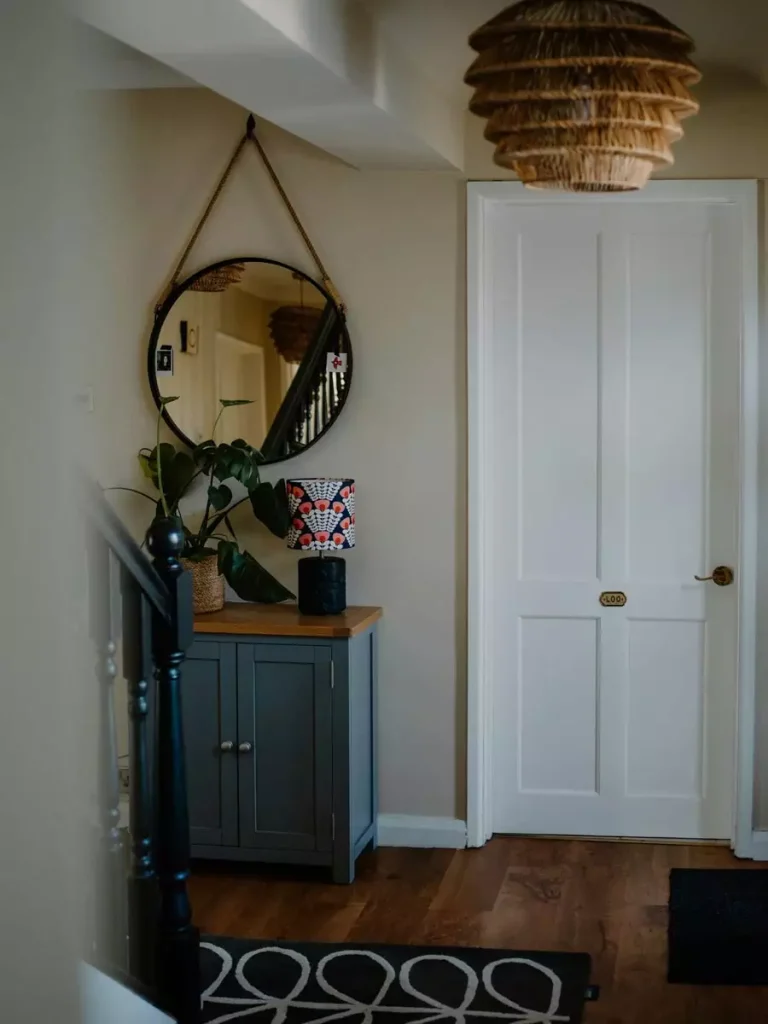 Small entryway with a strap hung round mirror above a blue cabinet and a woven pendant light