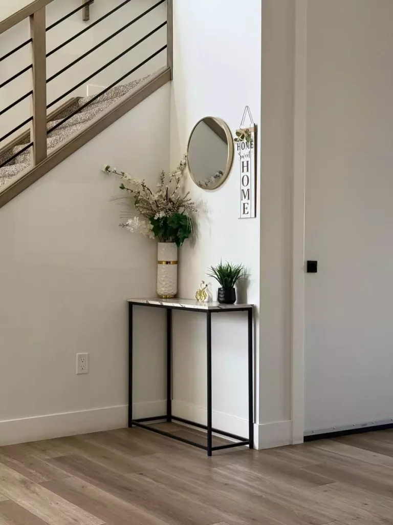 Small round mirror and vertical home sign above a narrow black console with flowers beside the staircase