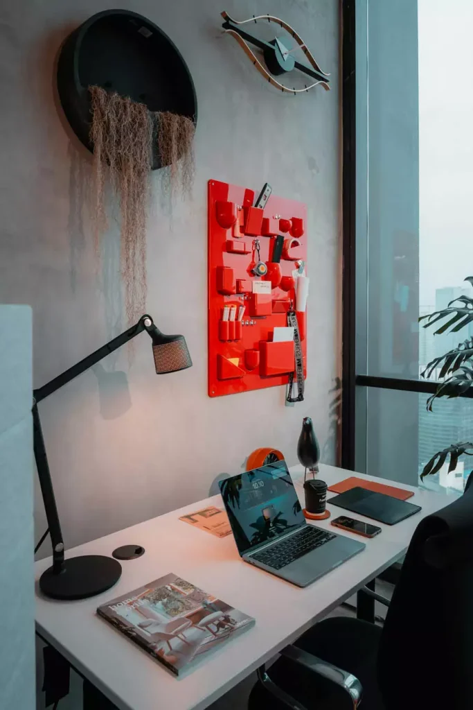 Study Desk Beneath A Red Wall Organizer A Round Decoration With Hanging Strands And An Eye Shaped Wall Ornament