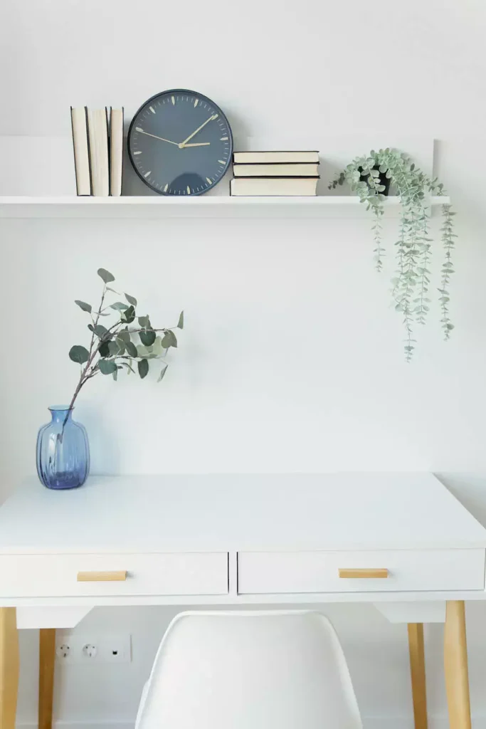 Study Desk Beneath A White Floating Shelf Holding Books A Dark Clock And A Trailing Plant