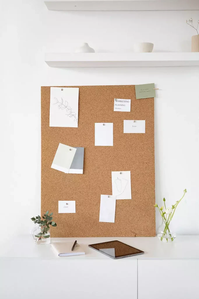 Study Wall Decor Featuring A Corkboard With Pinned Notes And Sketches Leaning On A Cabinet Beneath White Shelves
