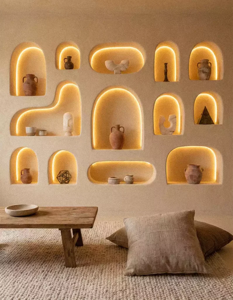 Textured beige wall with illuminated irregular niches displaying pottery and small sculptures