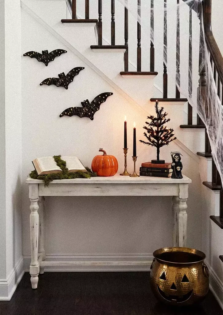 Three black bats above a white console table beneath stairs draped with cobwebs