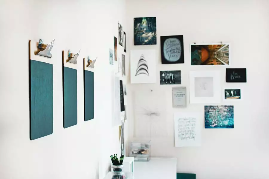Three teal clipboards hang beside a corner wall collage of photographs, art prints, and handwritten cards above a white desk